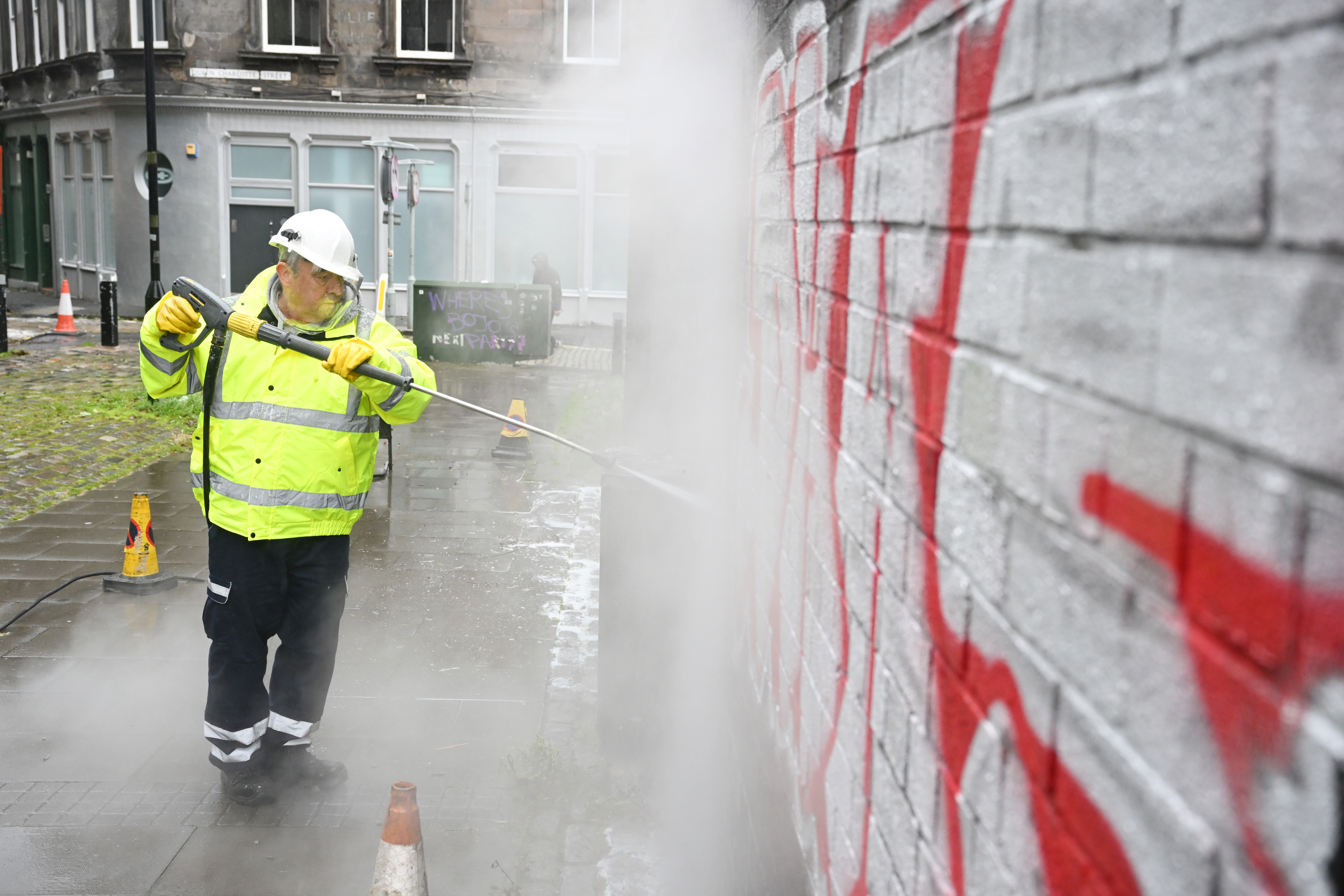 Photograph showing a wall covered in graffiti on the right and a man on the right cleaning the graffiti with a power washer